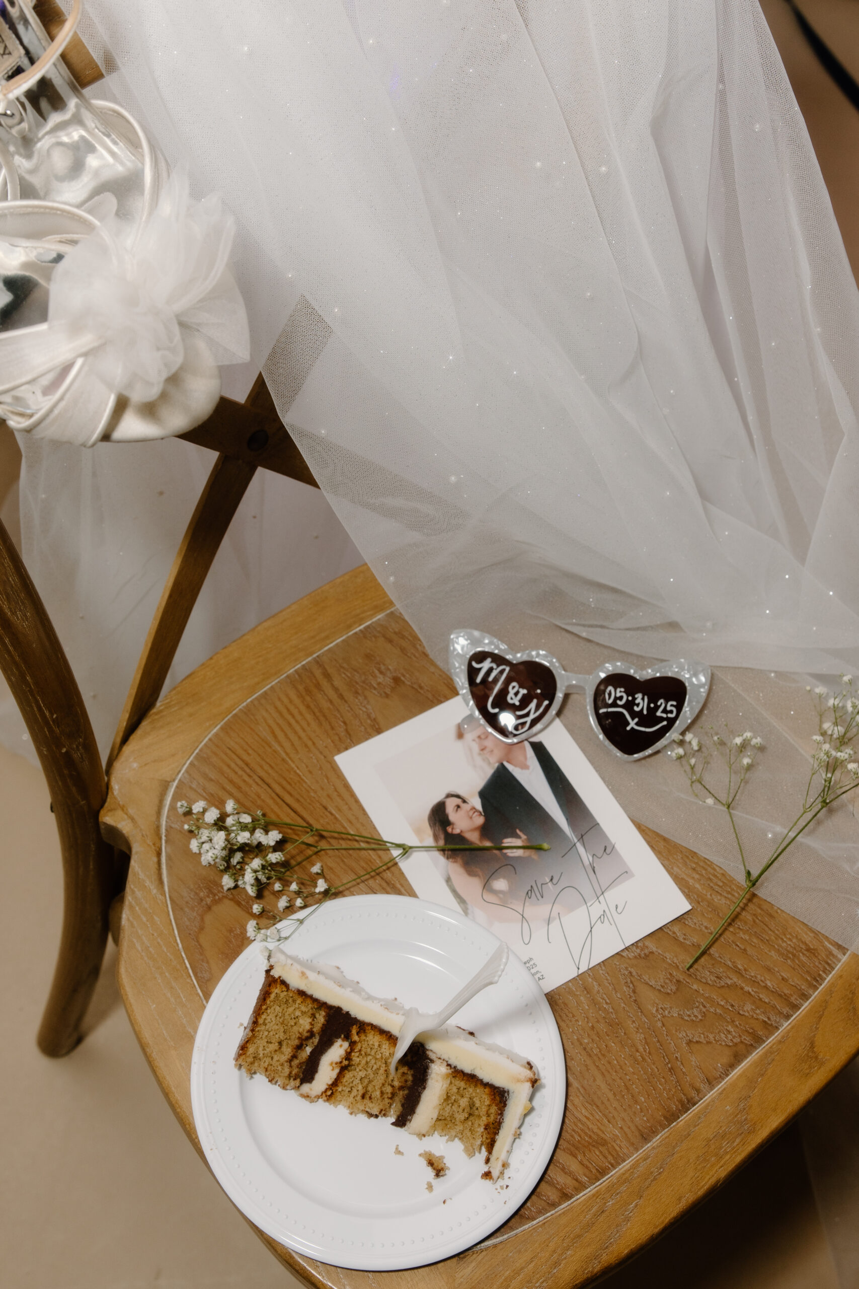 Wedding detail flat lay featuring heart-shaped sunglasses with the couple’s initials and date, a slice of cake, baby’s breath flowers, and a printed photo.