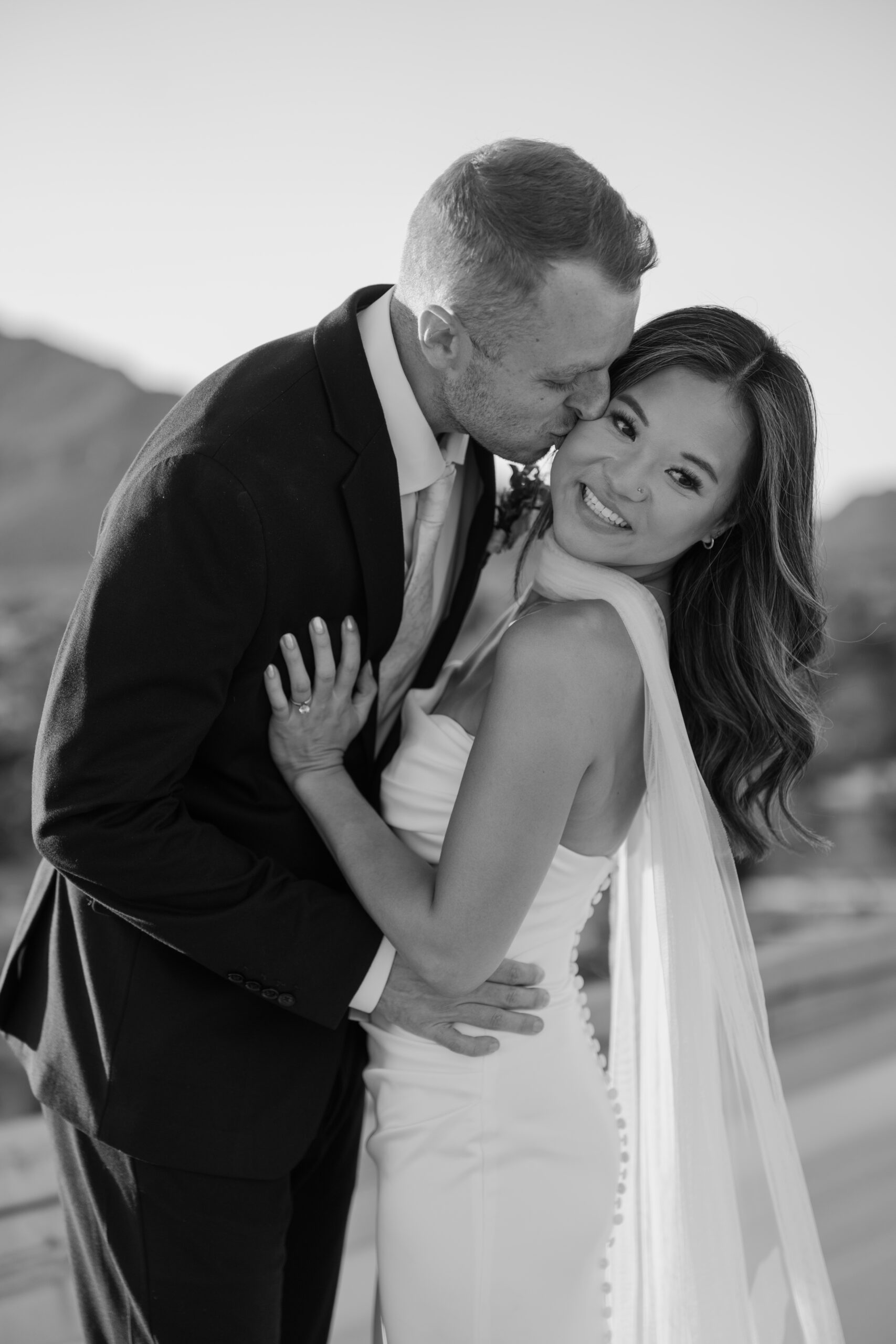 Black and white portrait of groom kissing bride’s cheek while she smiles, holding onto him