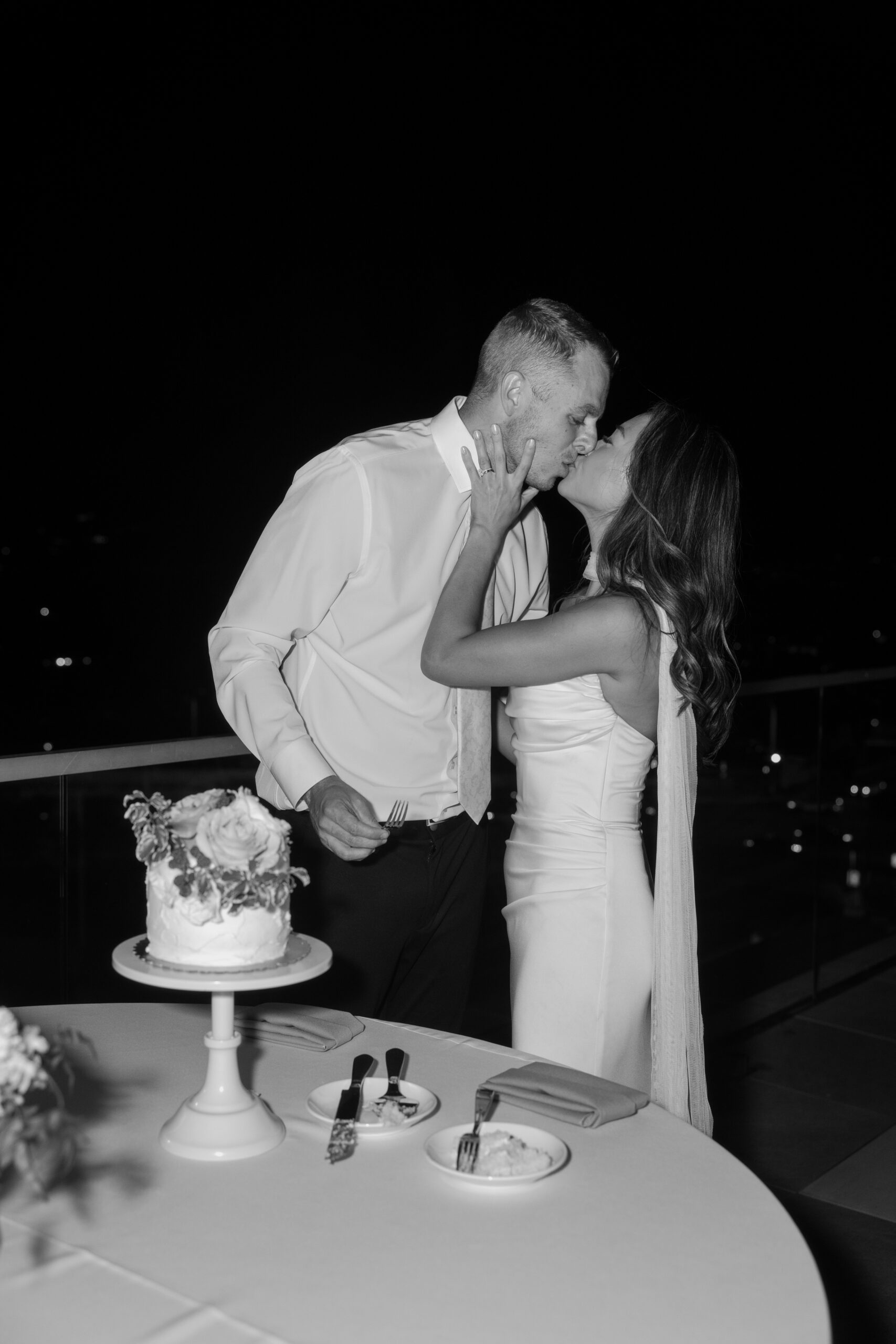 Black and white photo of bride and groom kissing while cutting their wedding cake at a reception table