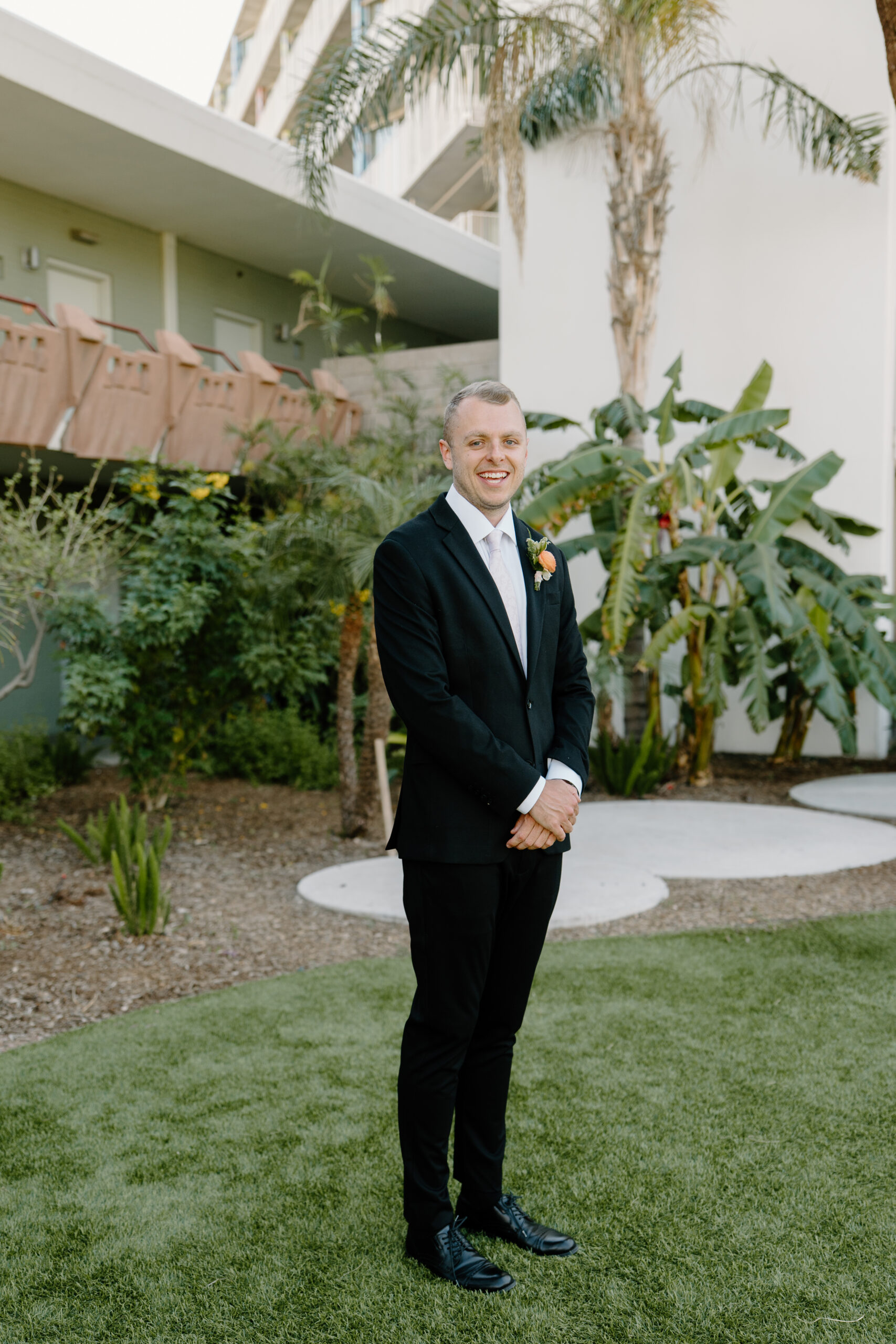 Groom standing in a black suit smiling in a garden setting with tropical greenery at Hotel Valley Ho