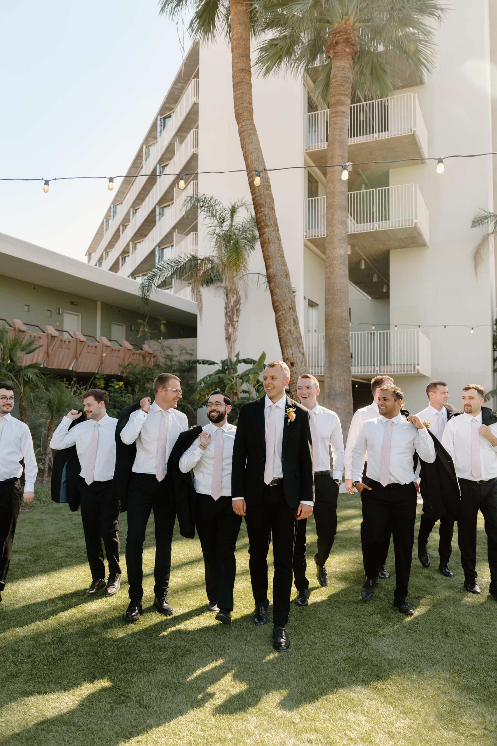 Groom and groomsmen walking together across the lawn in black suits with palm trees and mid-century architecture at Hotel Valley Ho