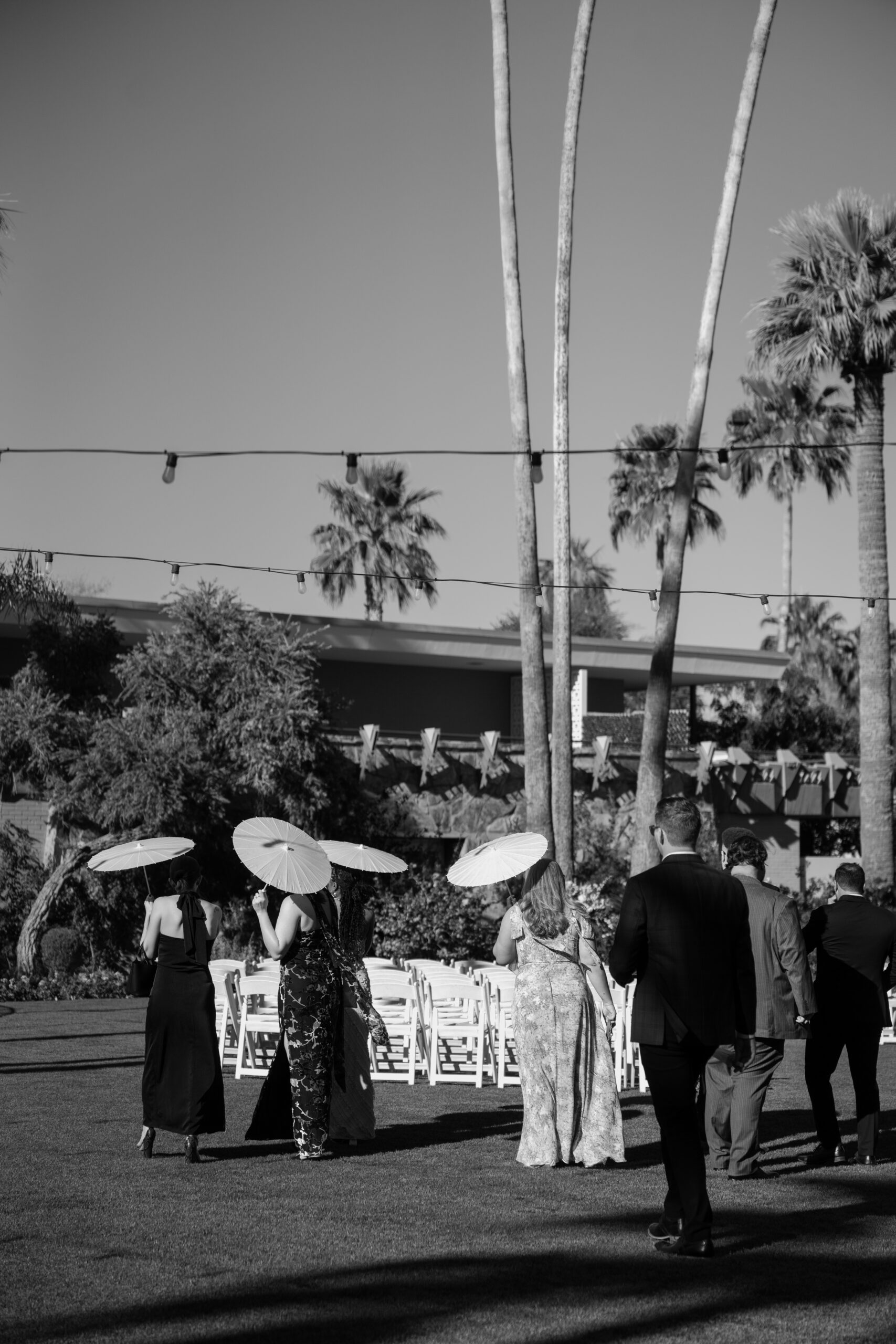 Black and white photo of guests walking toward the ceremony holding parasols under tall palm trees