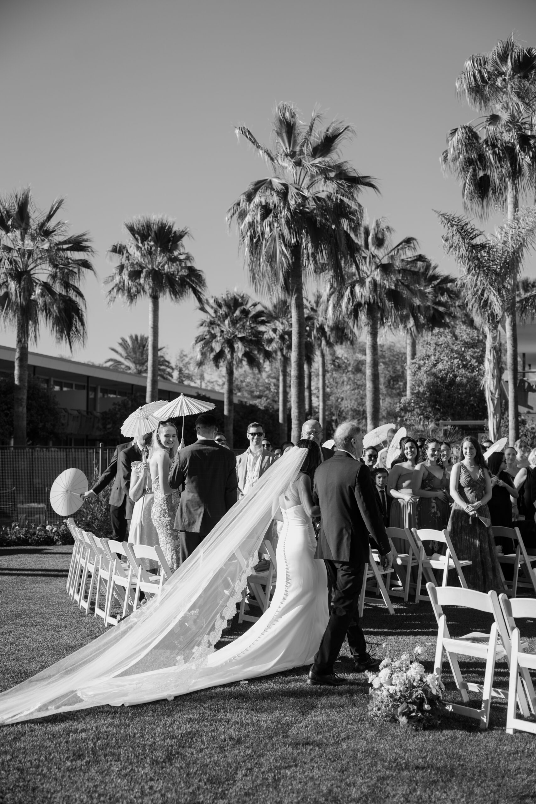 Black and white photo of bride walking down the aisle with a long flowing veil as guests look on during an outdoor ceremony