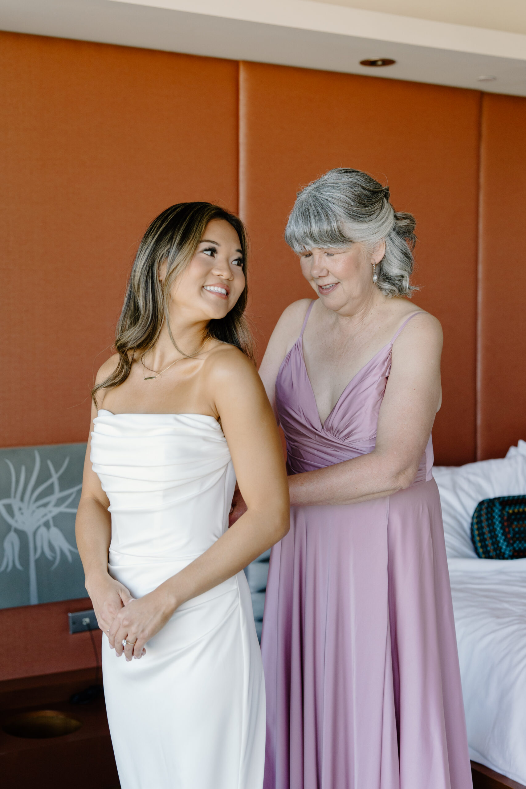 Bride smiling as an older woman helps zip up her wedding dress in a modern hotel room