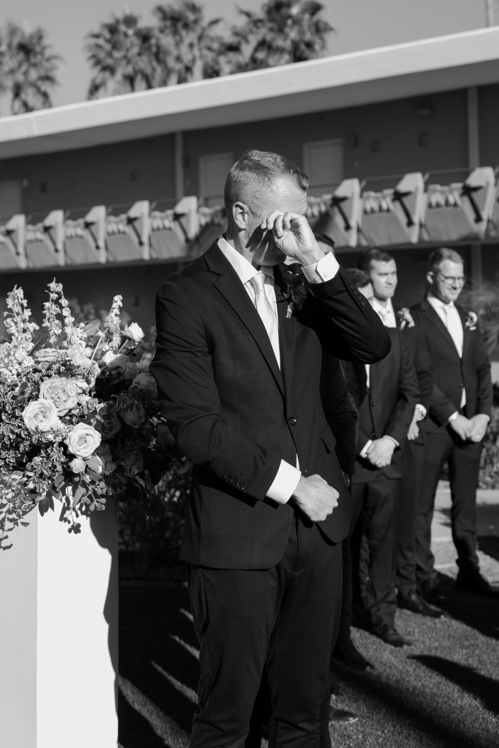Black and white photo of groom wiping away tears during the ceremony while groomsmen stand behind him
