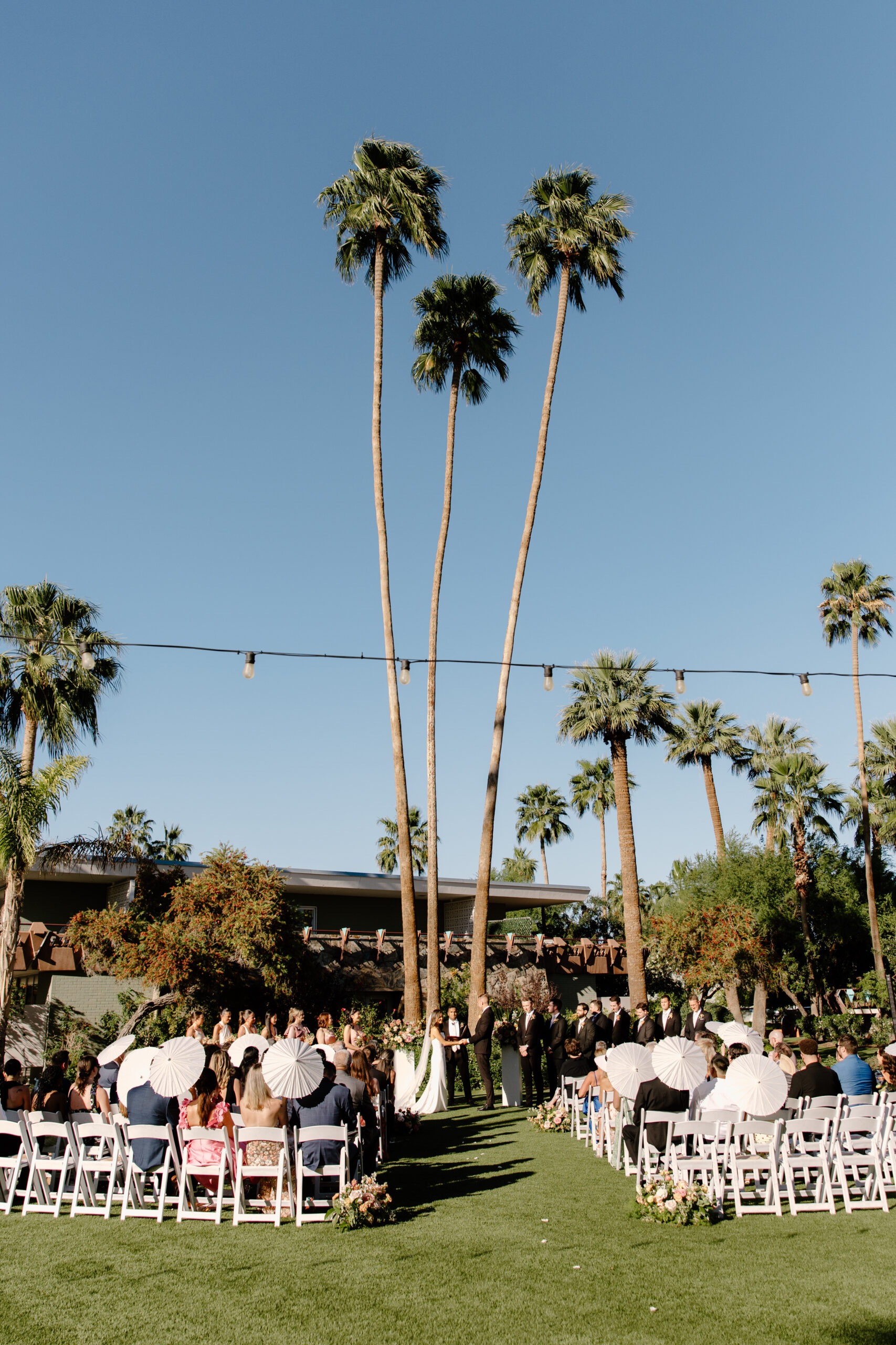 Wide view of an outdoor wedding ceremony with tall palm trees and guests seated in white chairs at Hotel Valley Ho