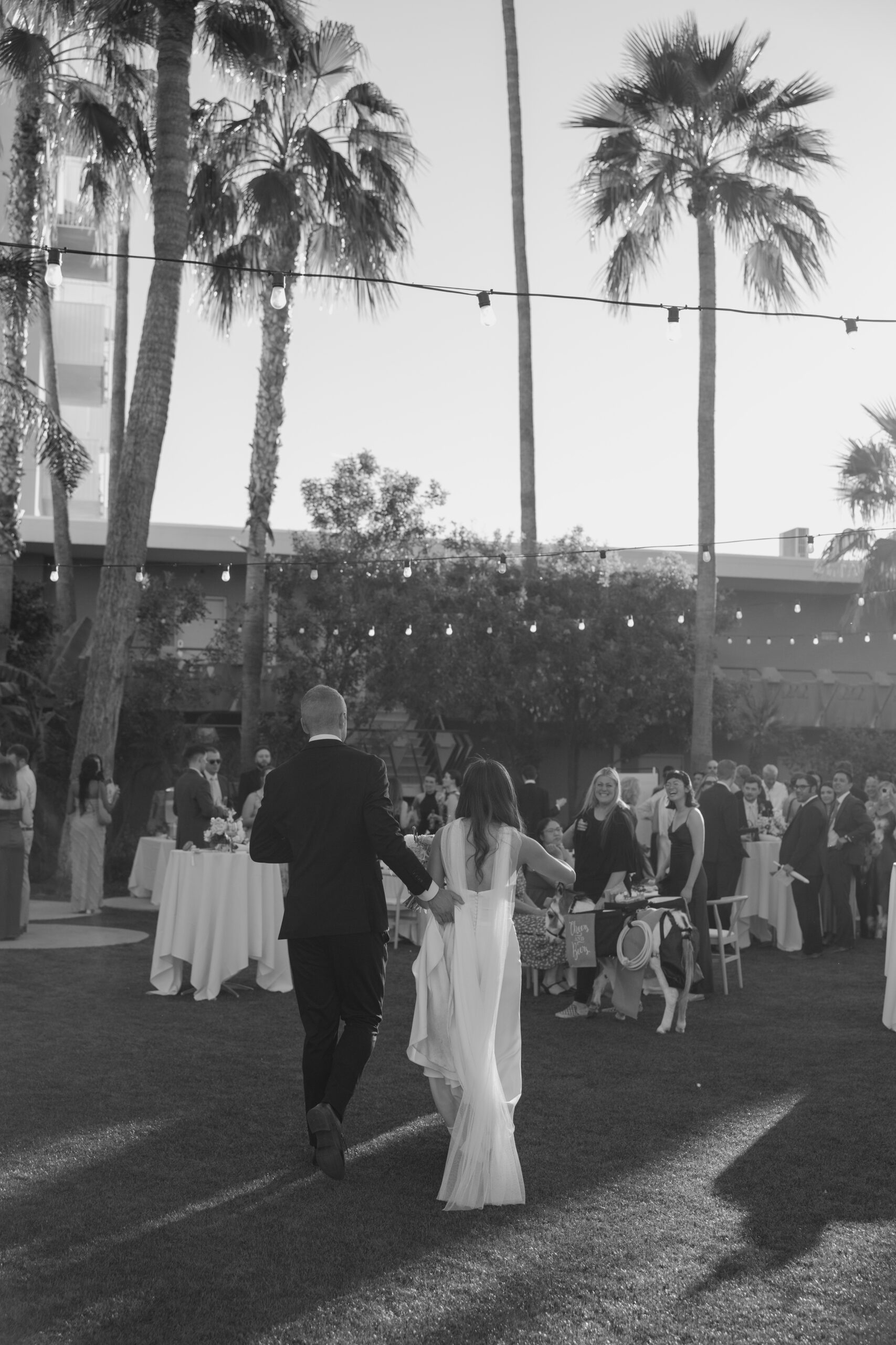 Black and white photo of newlyweds walking hand in hand into their reception space under string lights