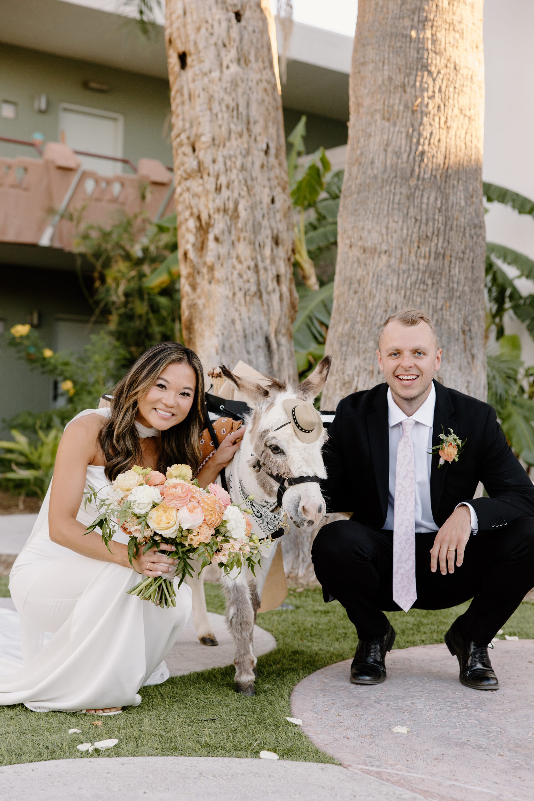 Bride and groom posing with a donkey wearing a small hat, smiling and holding a bouquet outdoors at Hotel Valley Ho