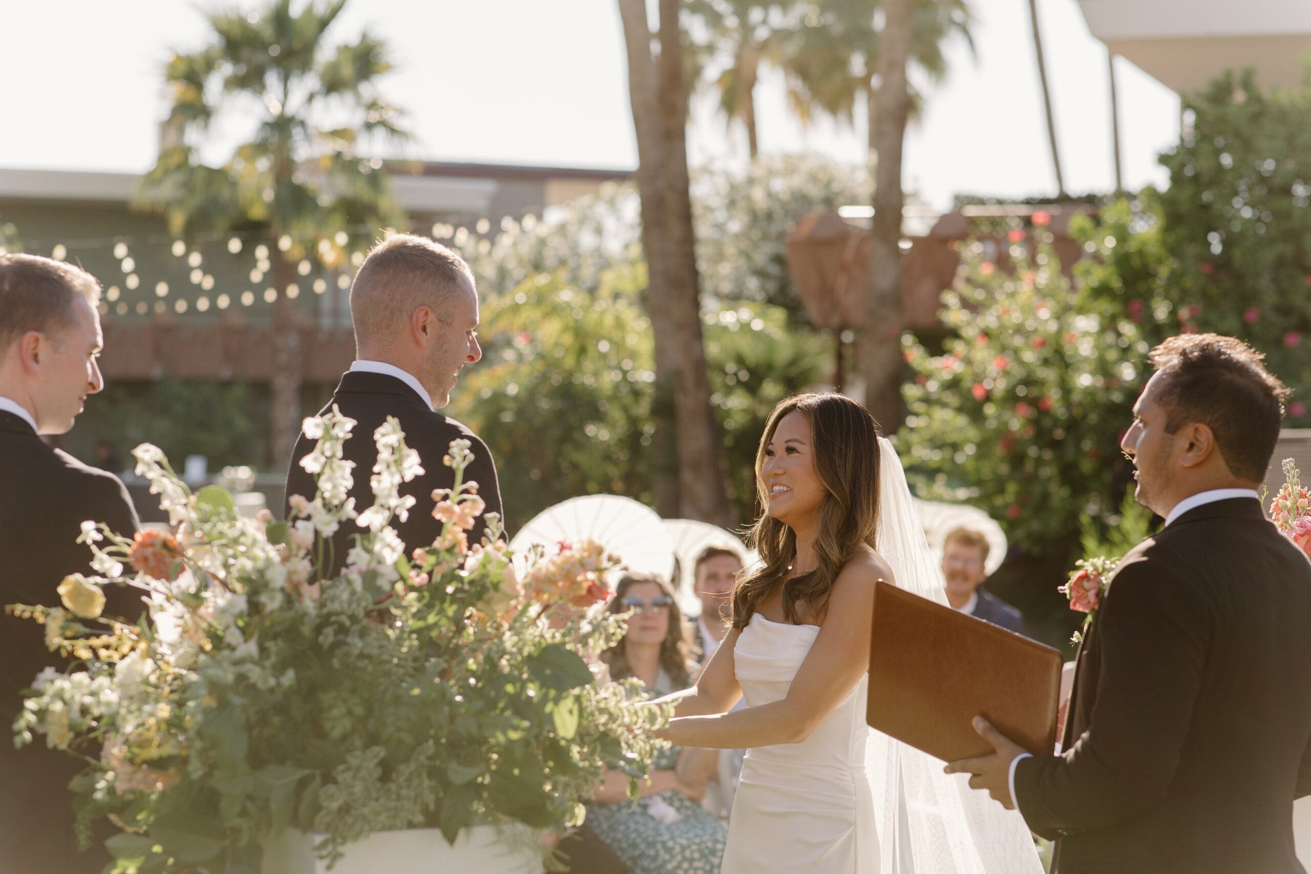 Bride and groom holding hands at the altar during a sunlit outdoor ceremony with guests in the background