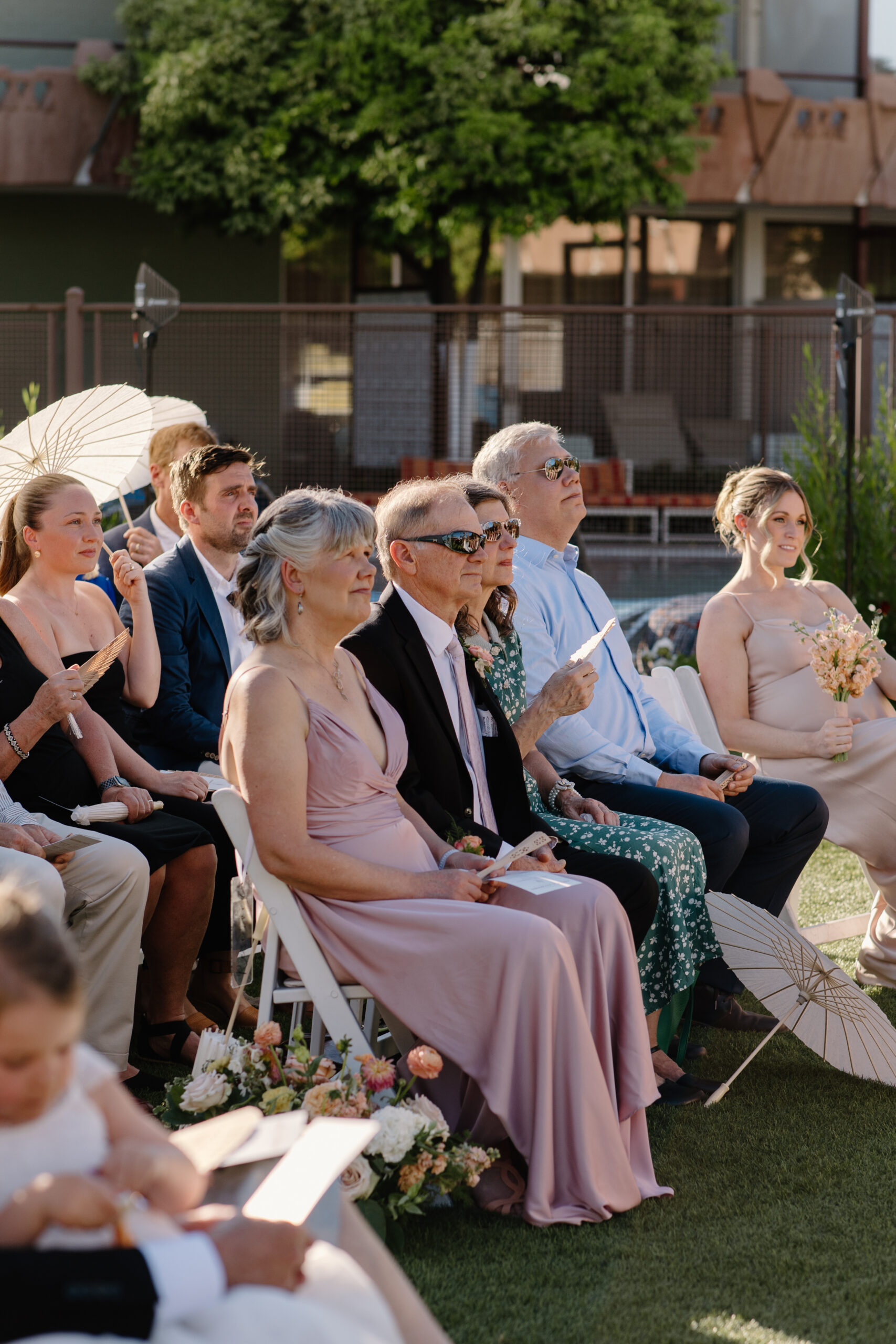 Wedding guests seated outdoors watching the ceremony, some holding parasols and programs in warm sunlight