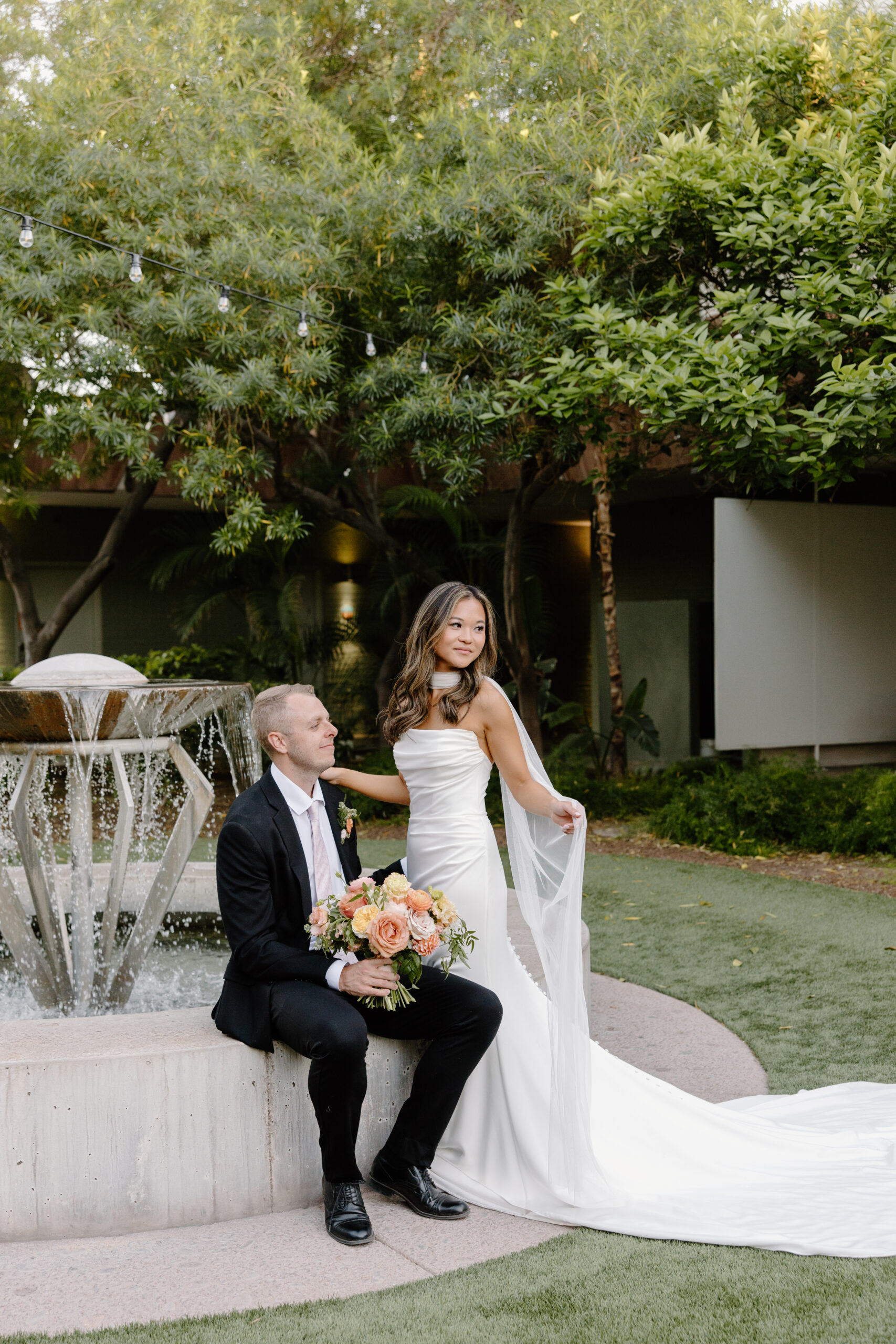 Bride and groom posing by a fountain surrounded by lush greenery at Hotel Valley Ho, with the bride holding her veil