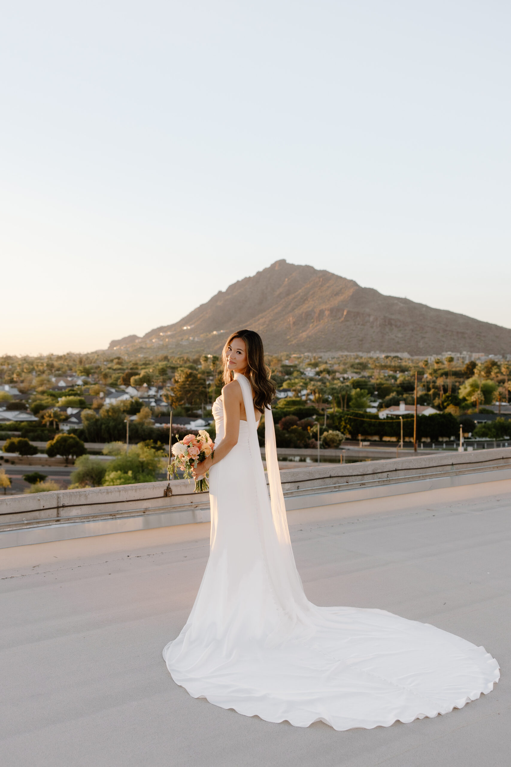 Bride in a sleek white gown holding a bouquet while standing on a rooftop with mountain views at sunset