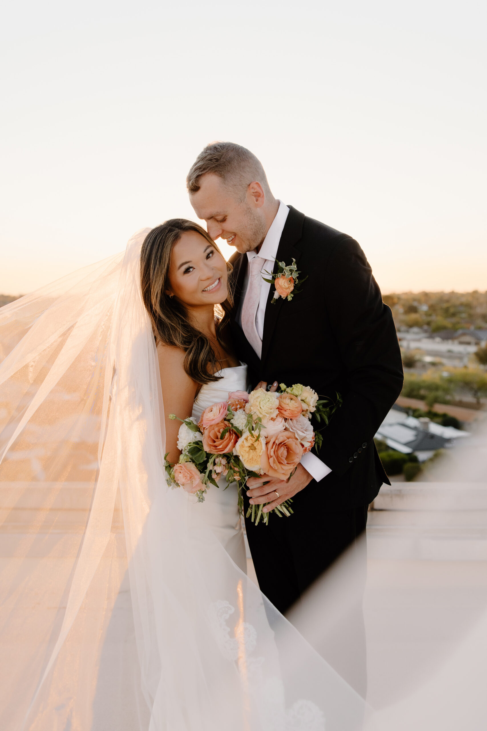 Romantic close-up of bride and groom embracing with soft veil movement and peach-toned bouquet during golden hour