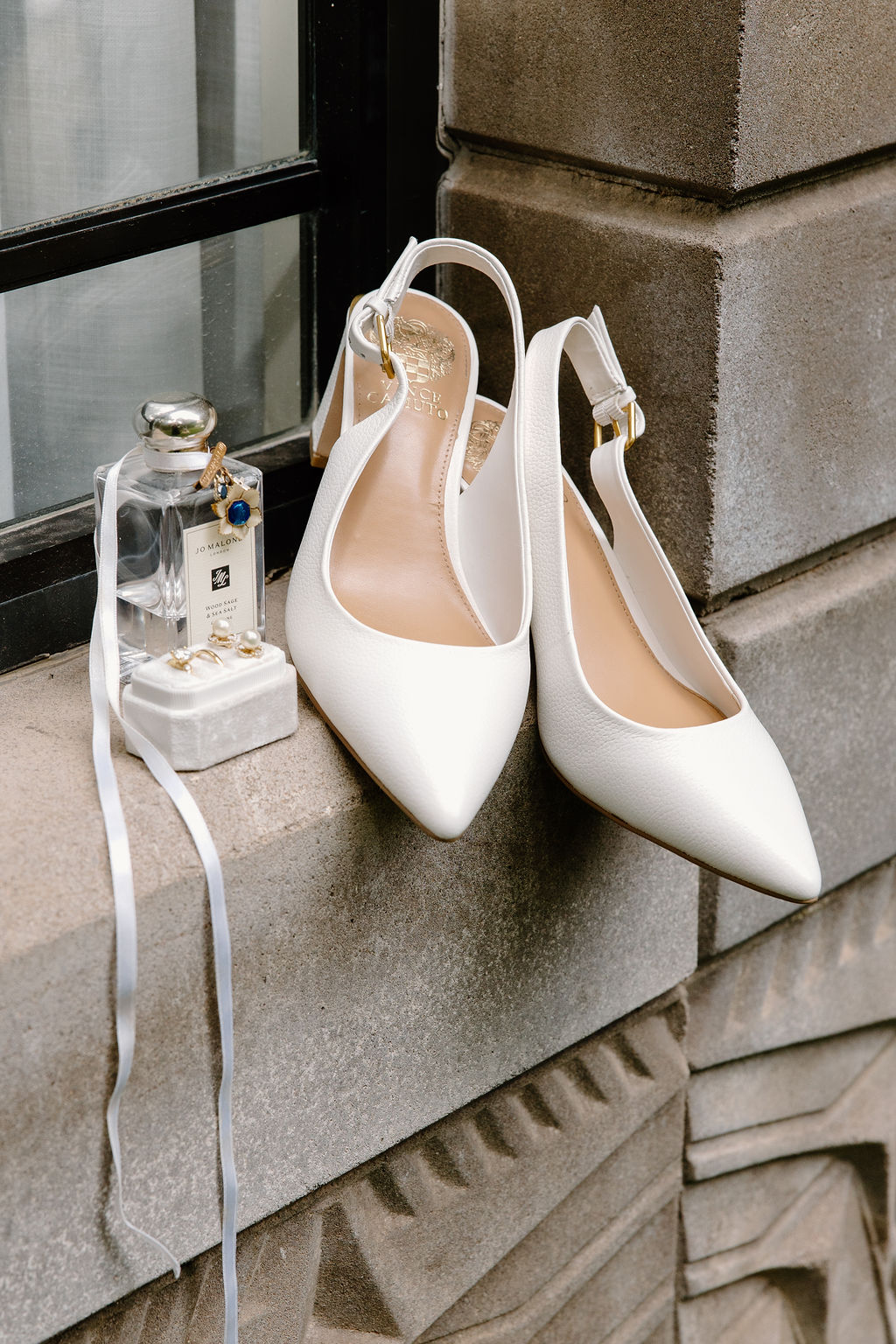 White bridal shoes styled with perfume, jewelry, and wedding accessories on a stone ledge.