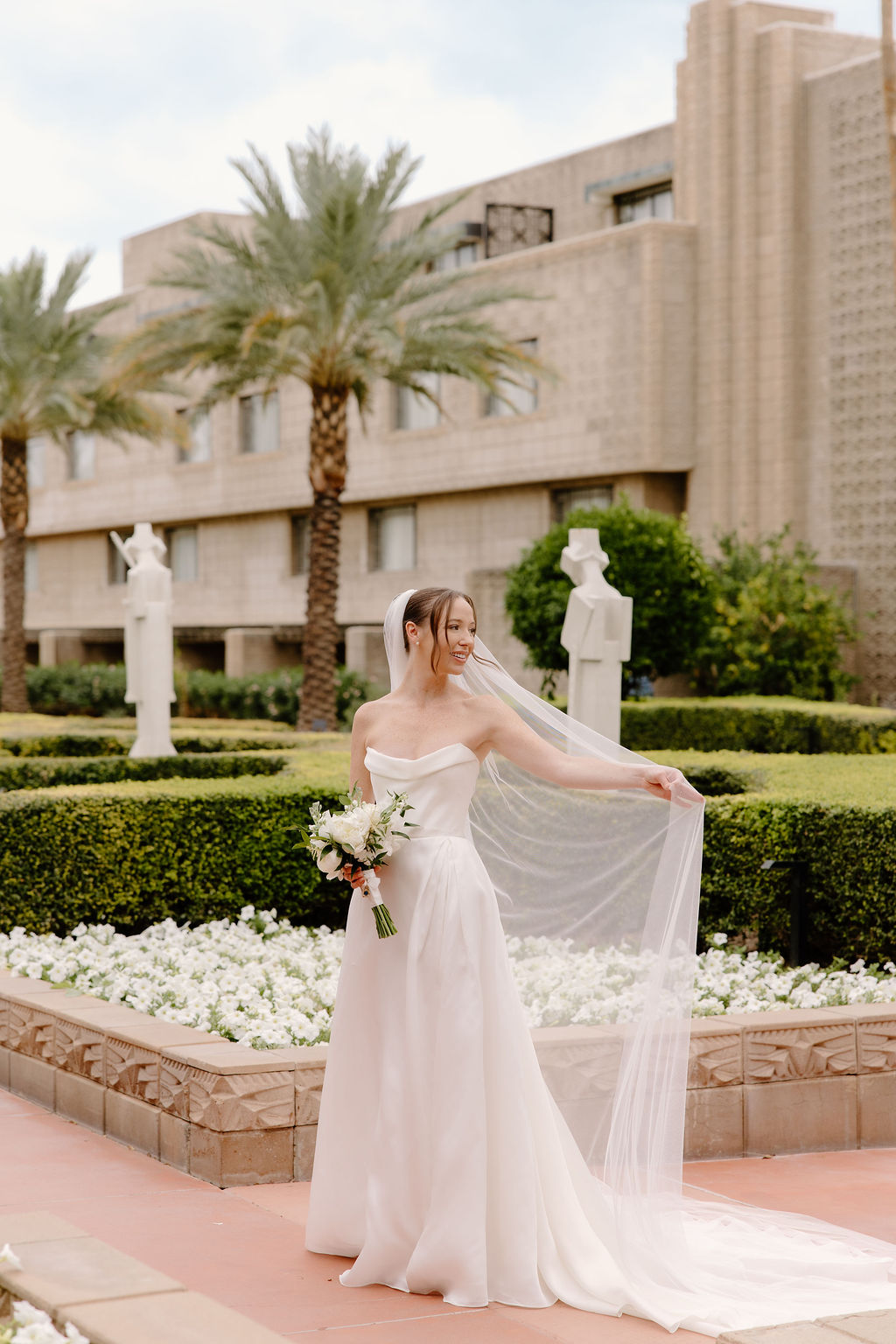 Bride posing with her long veil and white bouquet in a manicured garden.