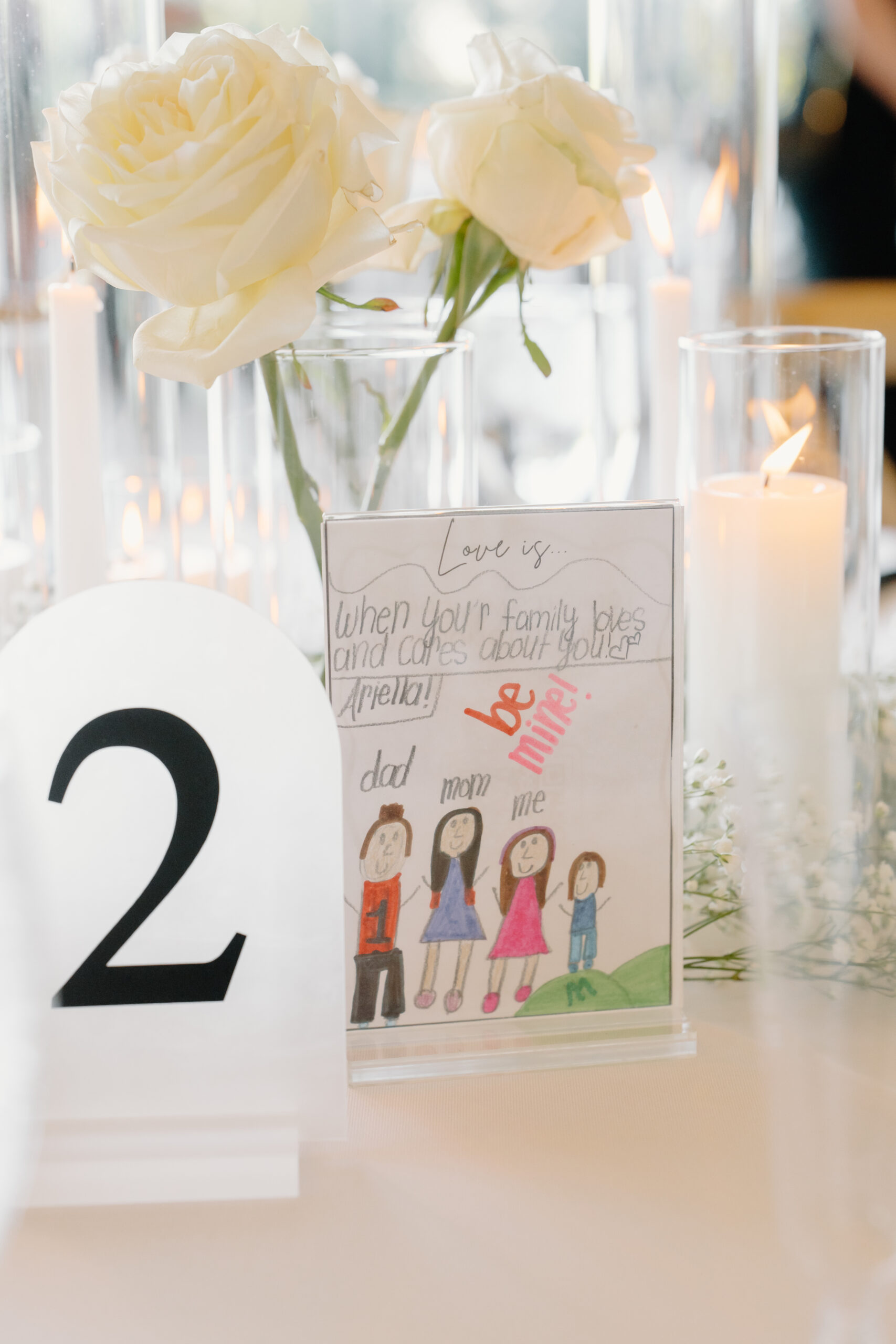 Wedding reception table detail with white roses, glowing candles, table number two, and a hand-drawn family note.