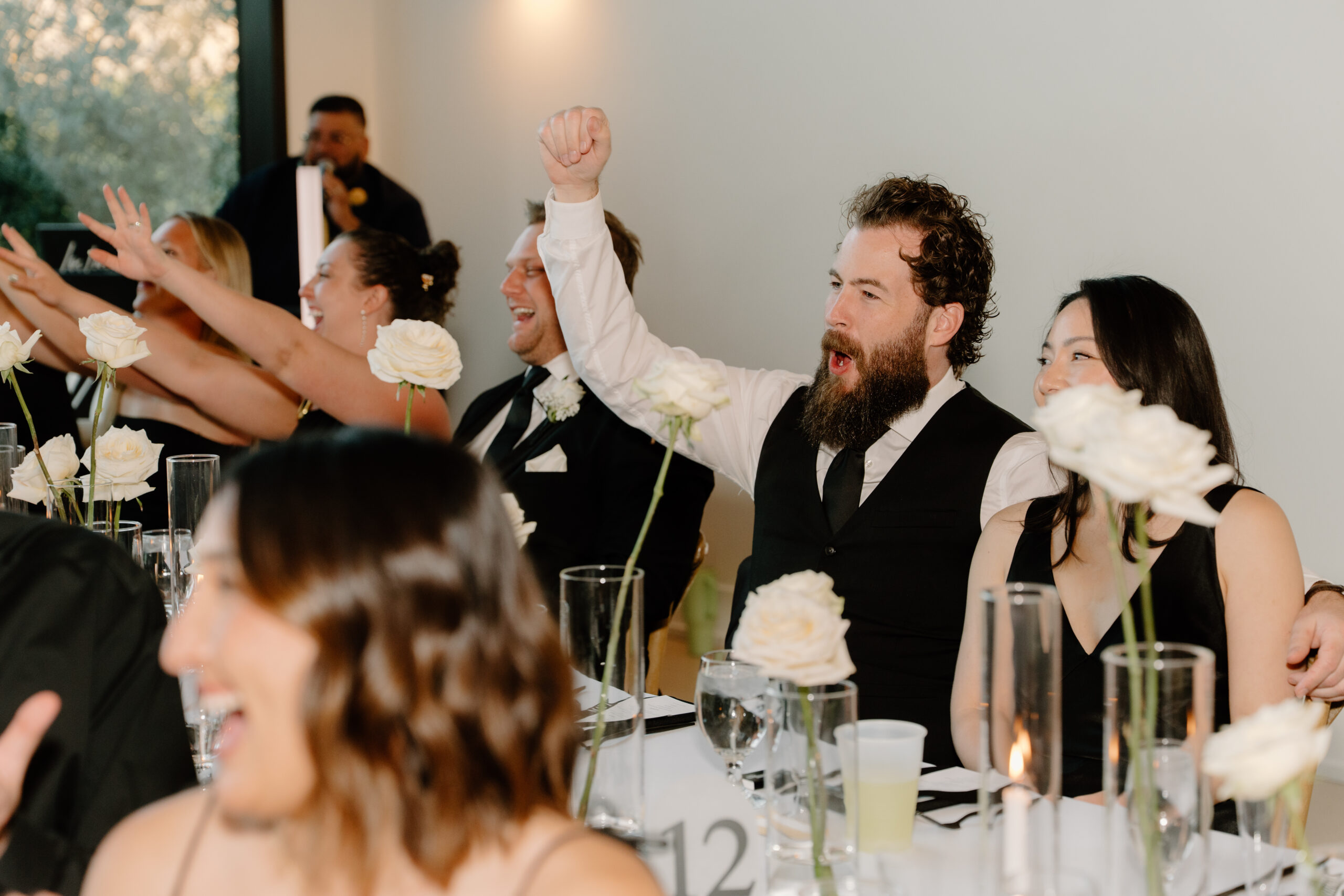 Wedding guests cheering during reception speeches at a candlelit table decorated with white roses, photographed by an Arizona wedding photographer.