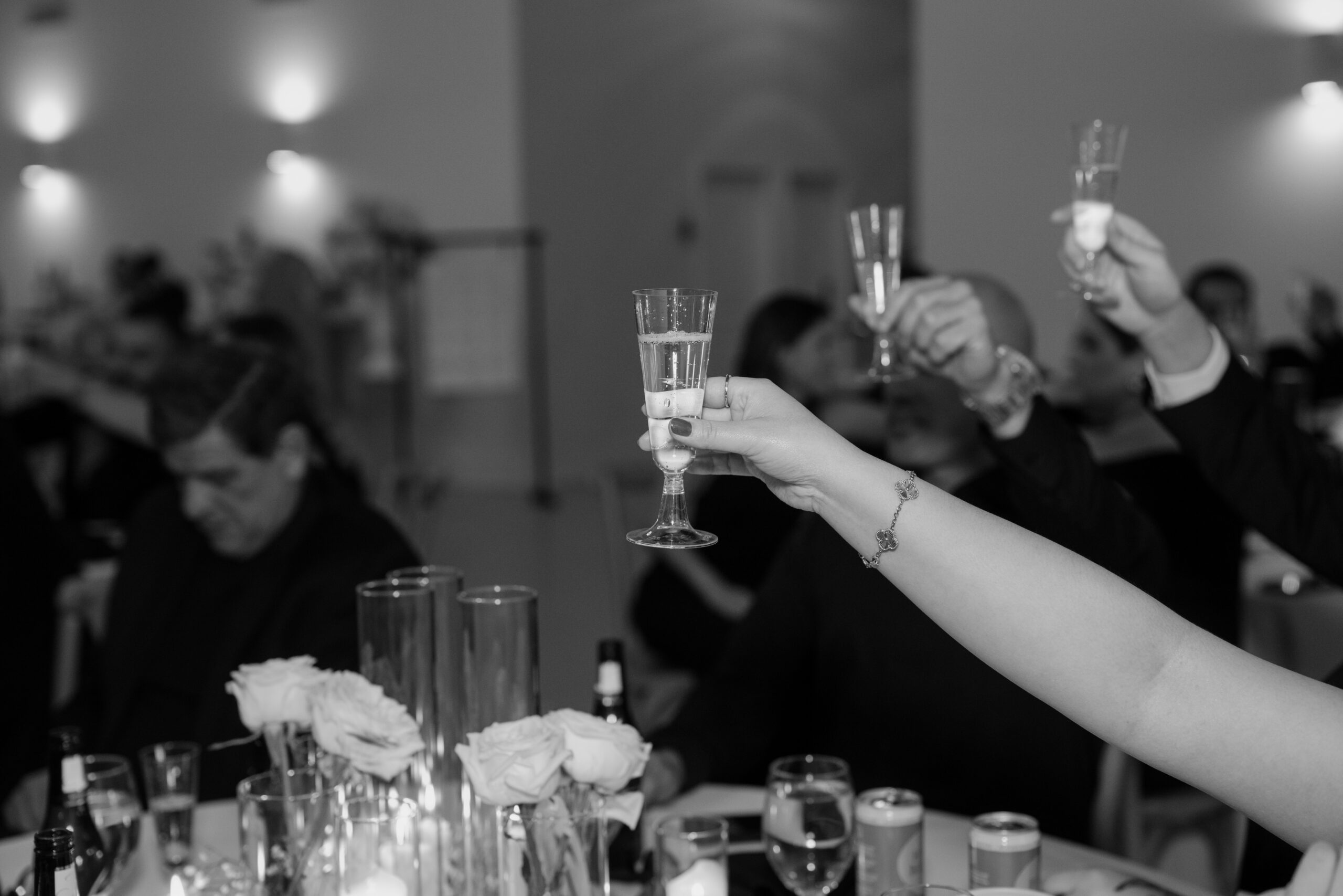 Black-and-white close-up of wedding guests raising champagne glasses for a celebratory reception toast.
