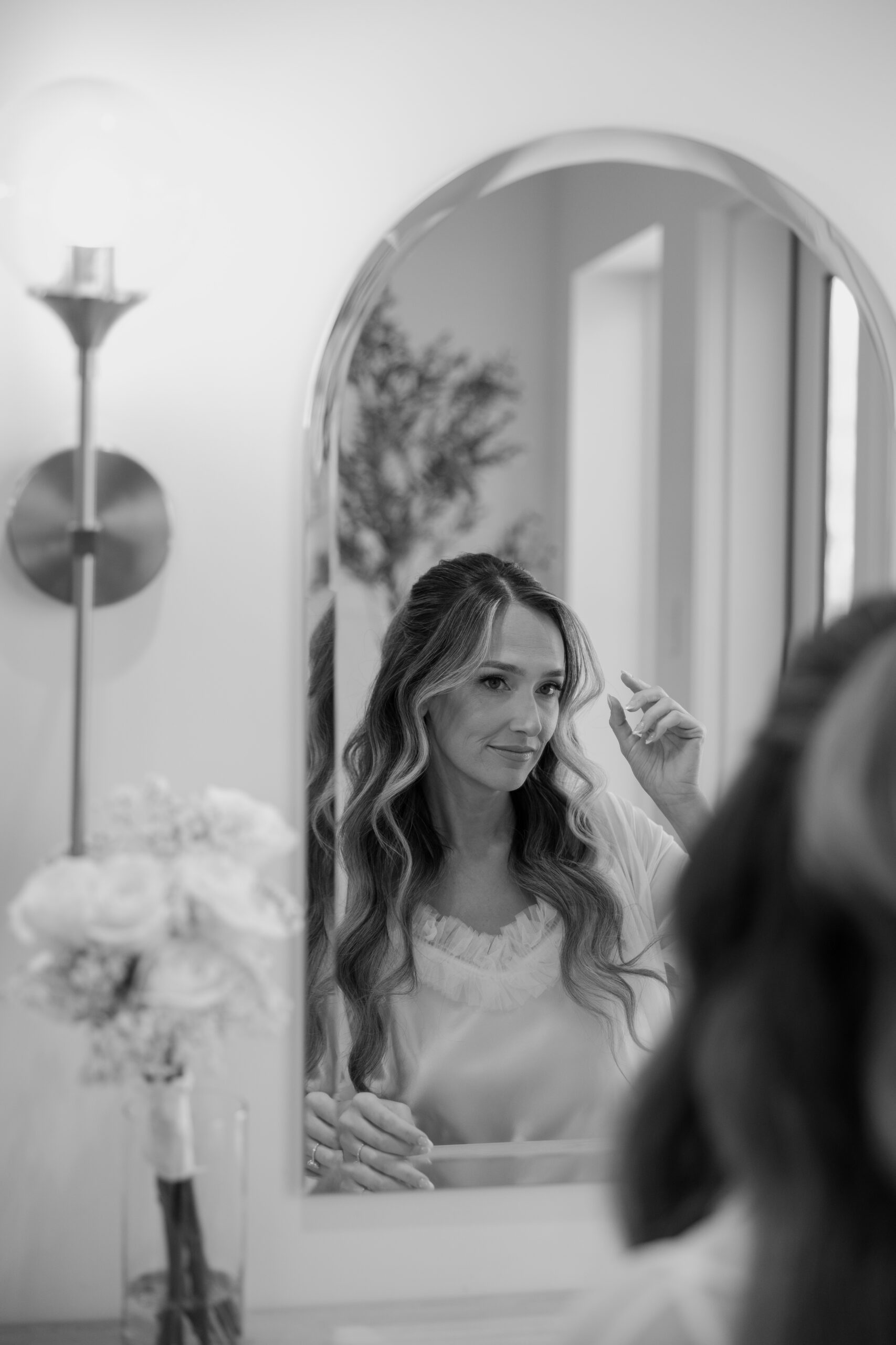 Black and white photo of the bride looking into a mirror while finishing her hair before the ceremony.
