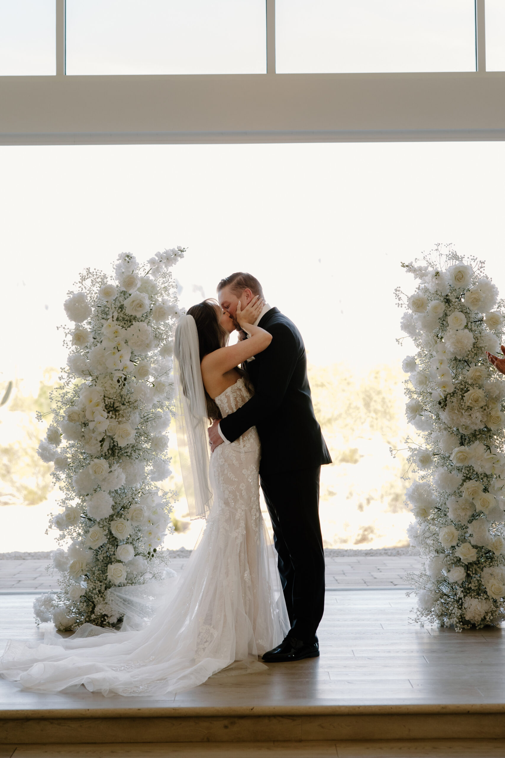 Newlyweds sharing their first kiss between tall white floral arrangements, a meaningful ceremony moment captured when choosing the right Arizona wedding photographer.
